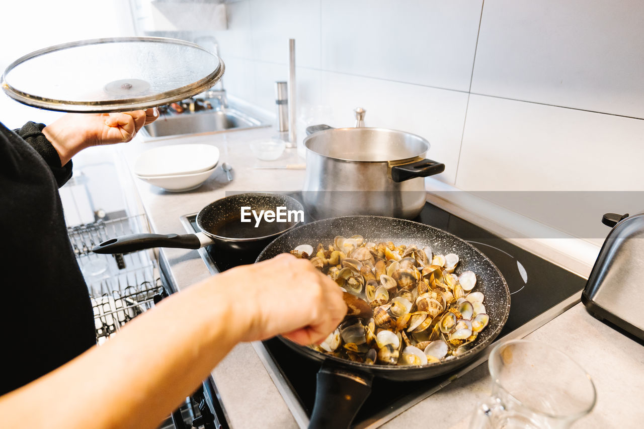 Heap of tasty mollusks with shells frying in hot black frying pan on electric cooker in light kitchen at home