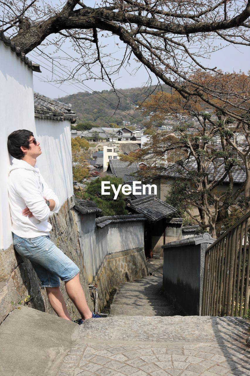 Side view of young man standing by tree against building