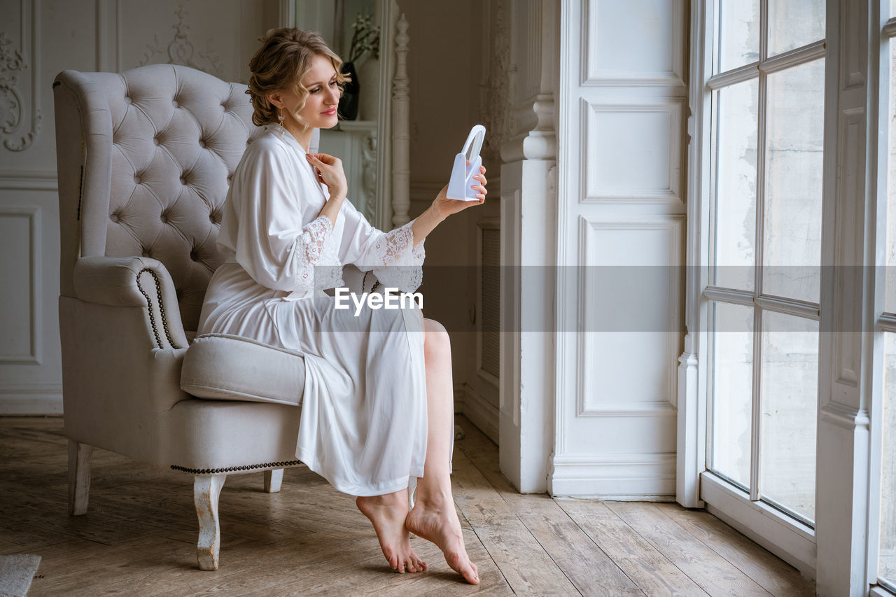 Charming woman in white robe applying body lotion while sitting in chair