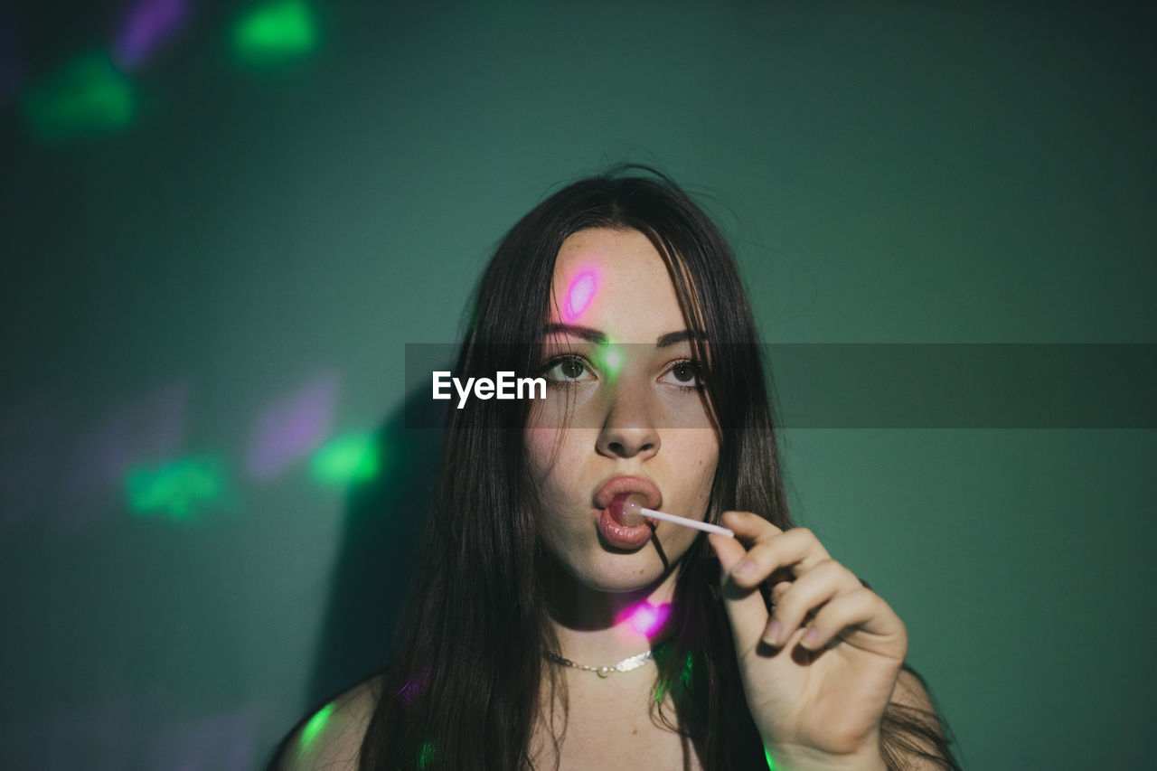 Young woman licking lollipop while lights falling on her against green background