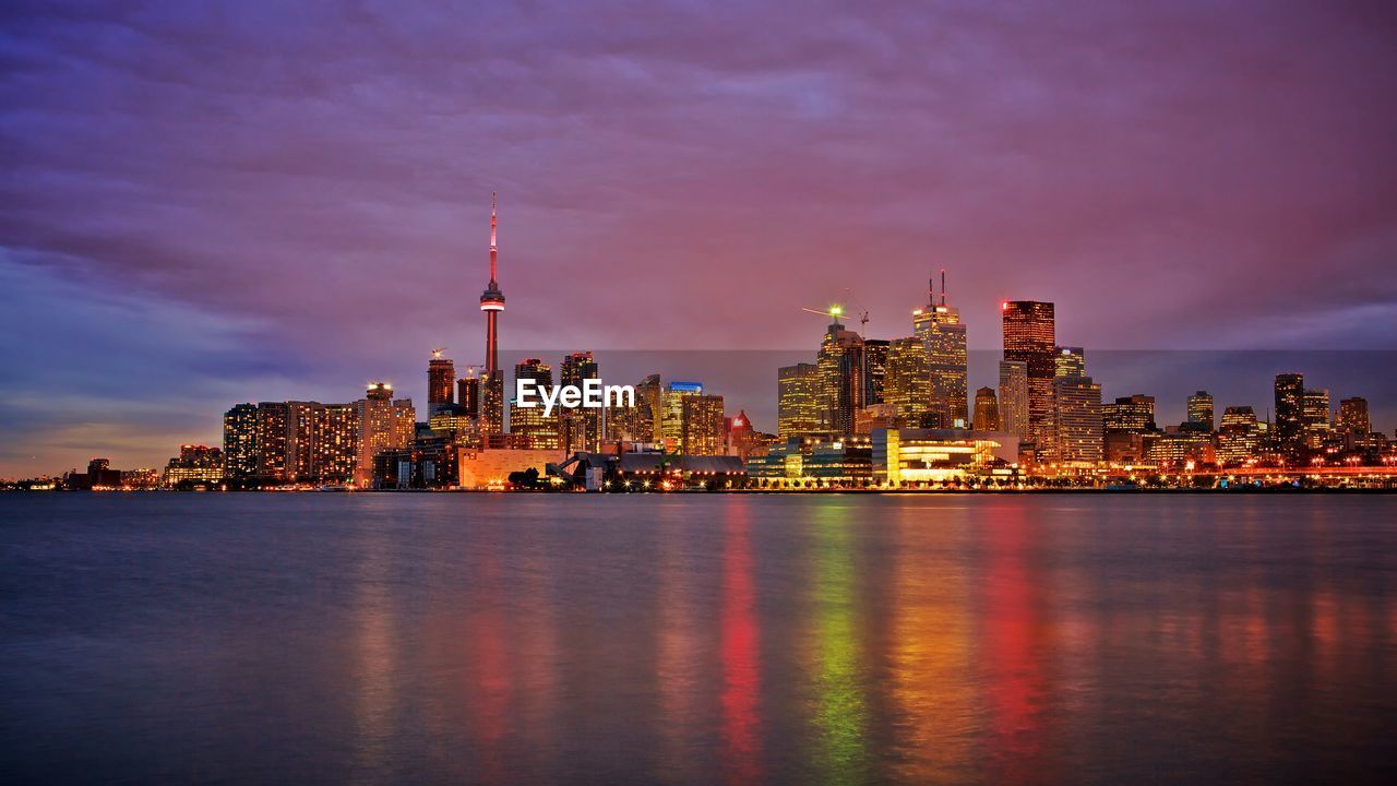 Sea by illuminated modern city skyline against sky during sunset