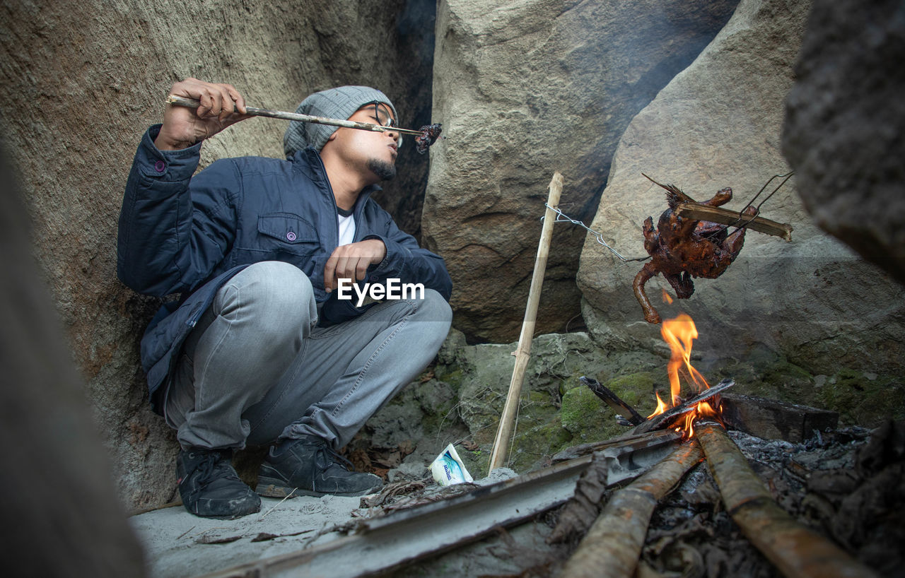 Man roasting meat over fire while sitting by rocks