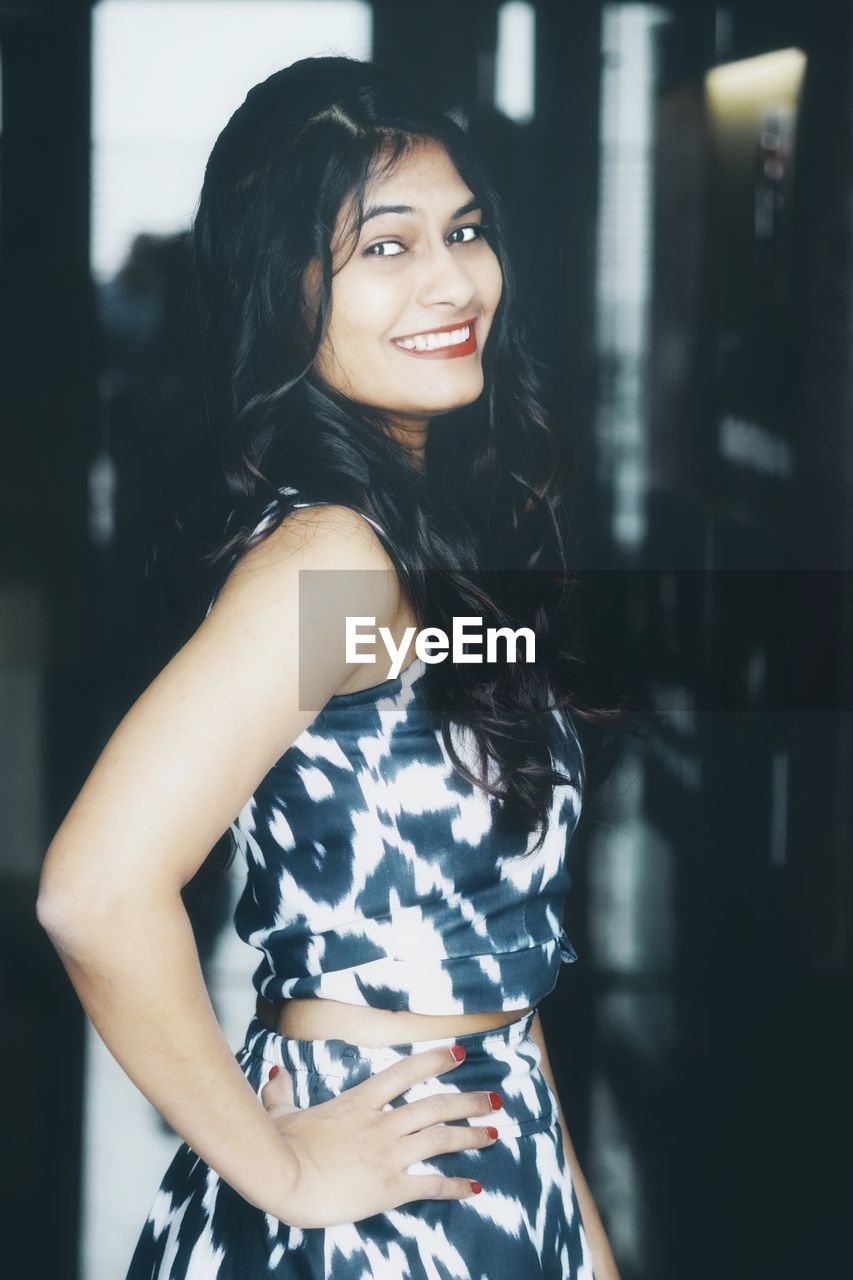 PORTRAIT OF A SMILING YOUNG WOMAN STANDING AT HOME