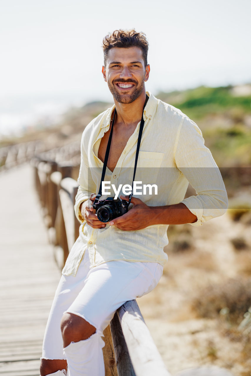 Portrait of smiling man holding camera sitting on railing