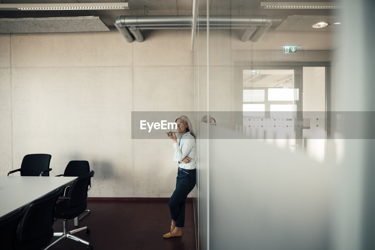 Businesswoman sending voicemail through smart phone leaning on glass wall in office