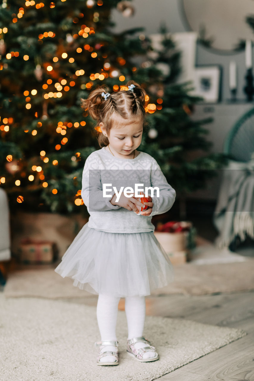 A beautiful little girl in a festive dress is standing at the christmas tree. 