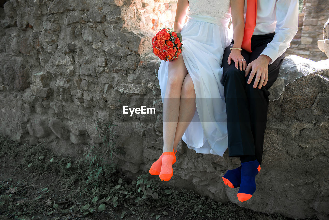 Low section of bride and groom sitting on stone wall