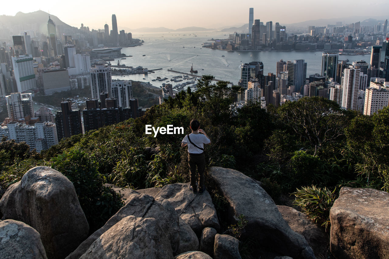 Rear view of man looking at city while standing by rocks against sky during sunset