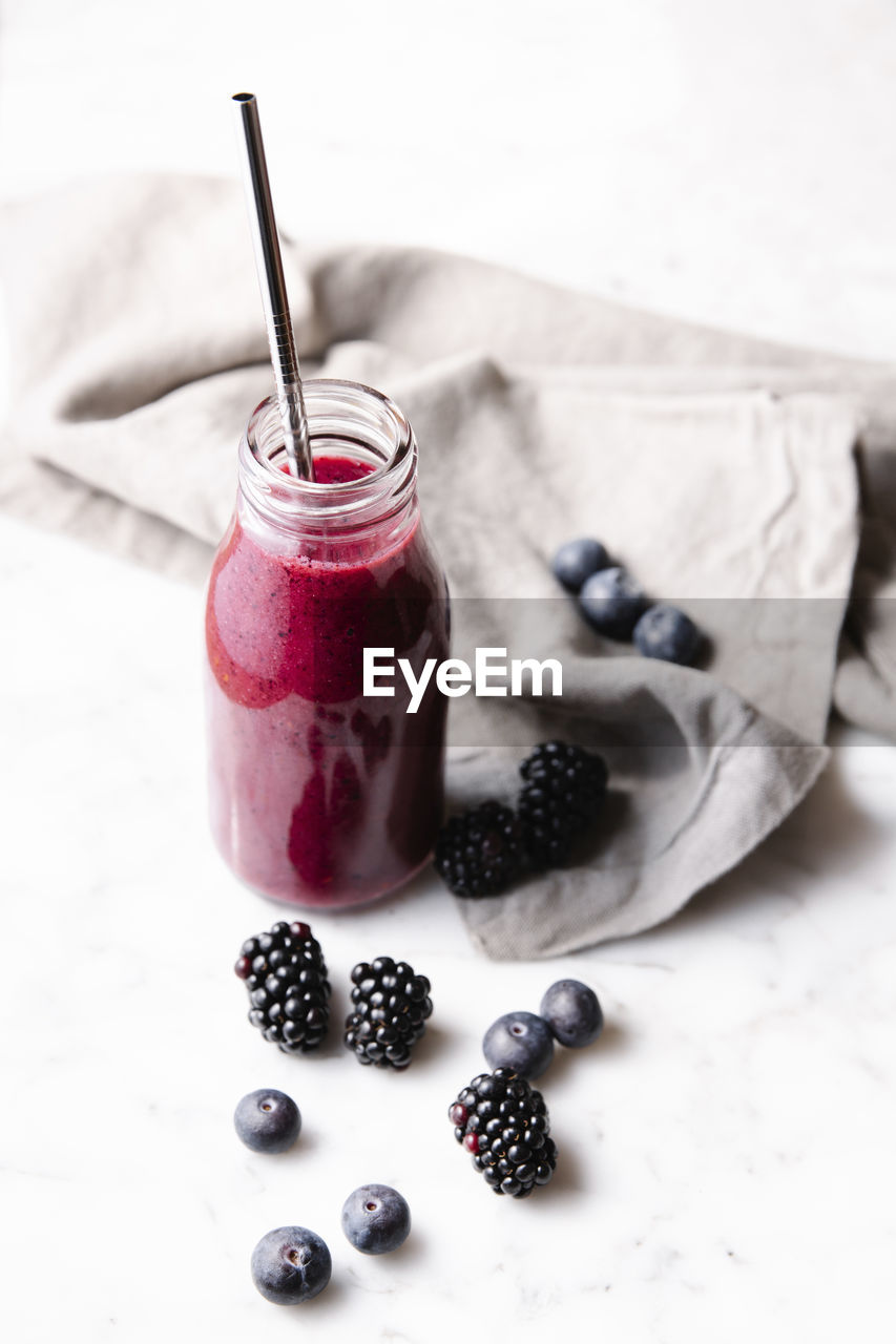 Glass bottle with fresh homemade berry smoothie and a reusable metal straw. some berries near the bottle on a marble surface. high angle vertical image.