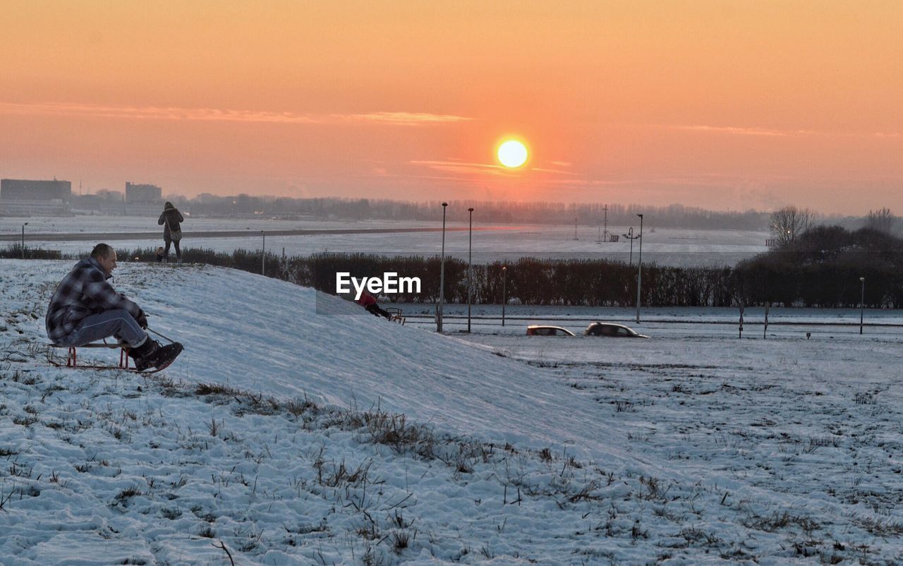 Side view full length of man tobogganing on snow covered field during sunset