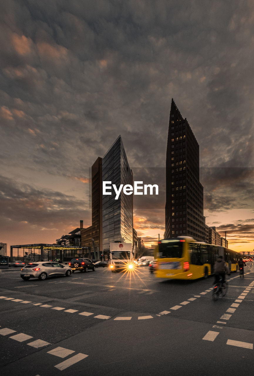 TRAFFIC ON ROAD BY BUILDINGS AGAINST SKY AT SUNSET