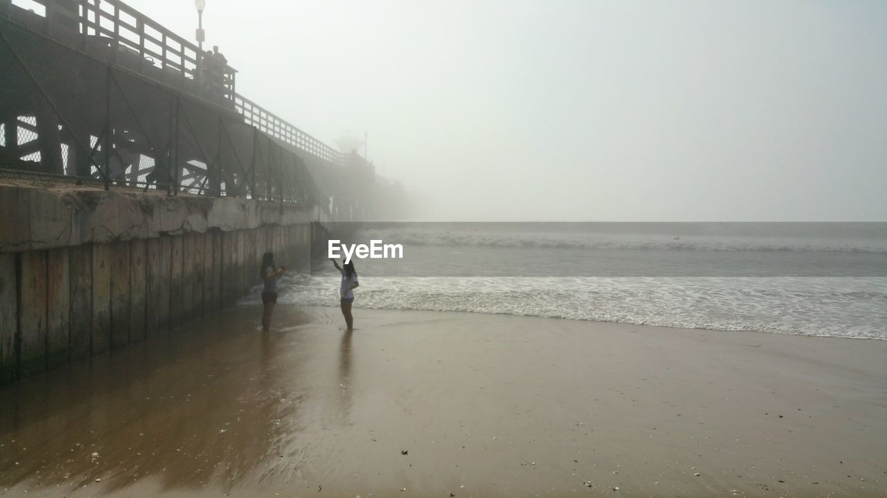 Women by pier at beach against sky during foggy weather