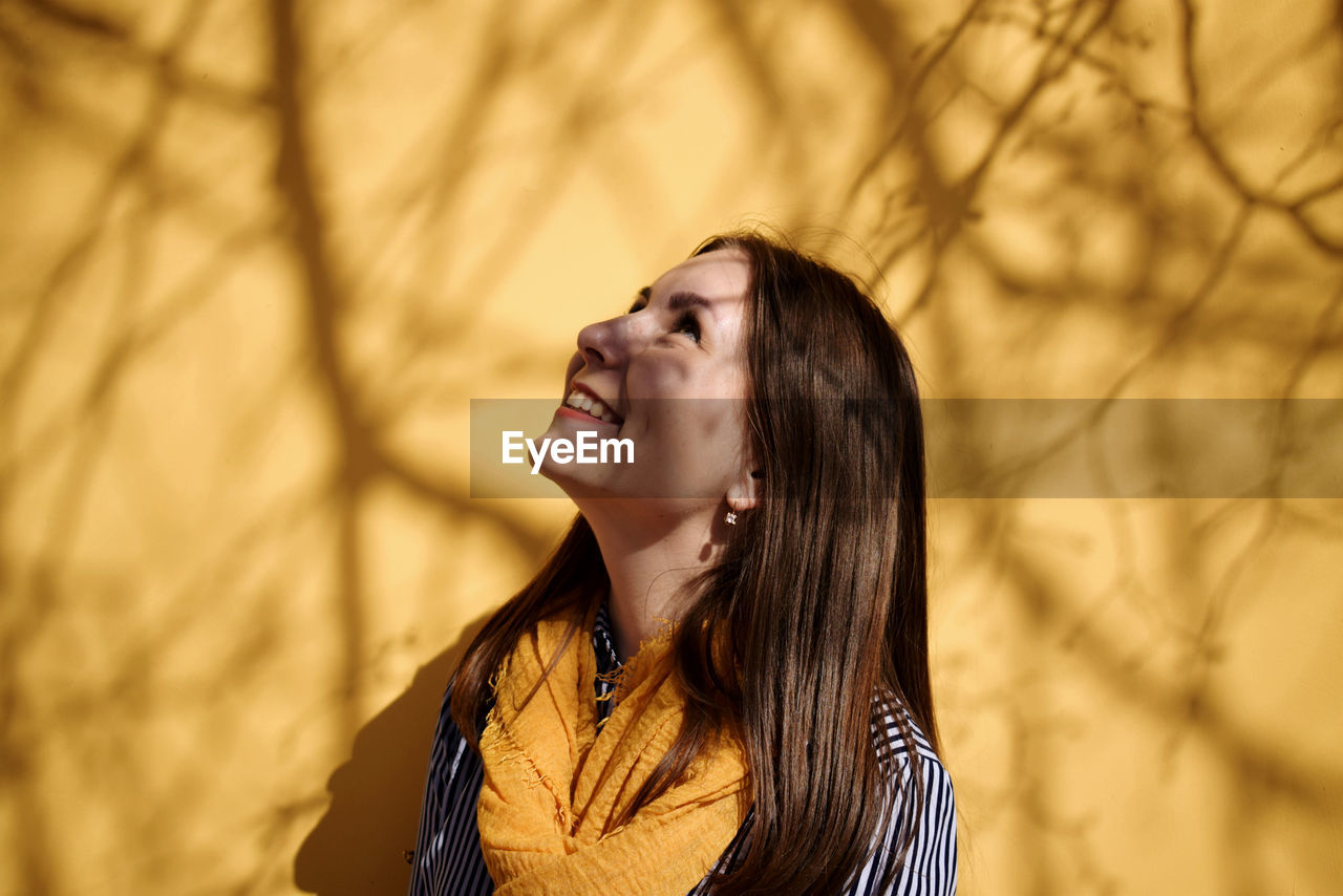 Smiling young woman looking up while standing against yellow wall