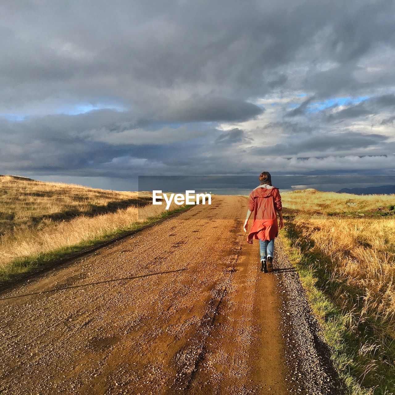 Rear view of young woman walking along dirt road in green field against cloudy sky