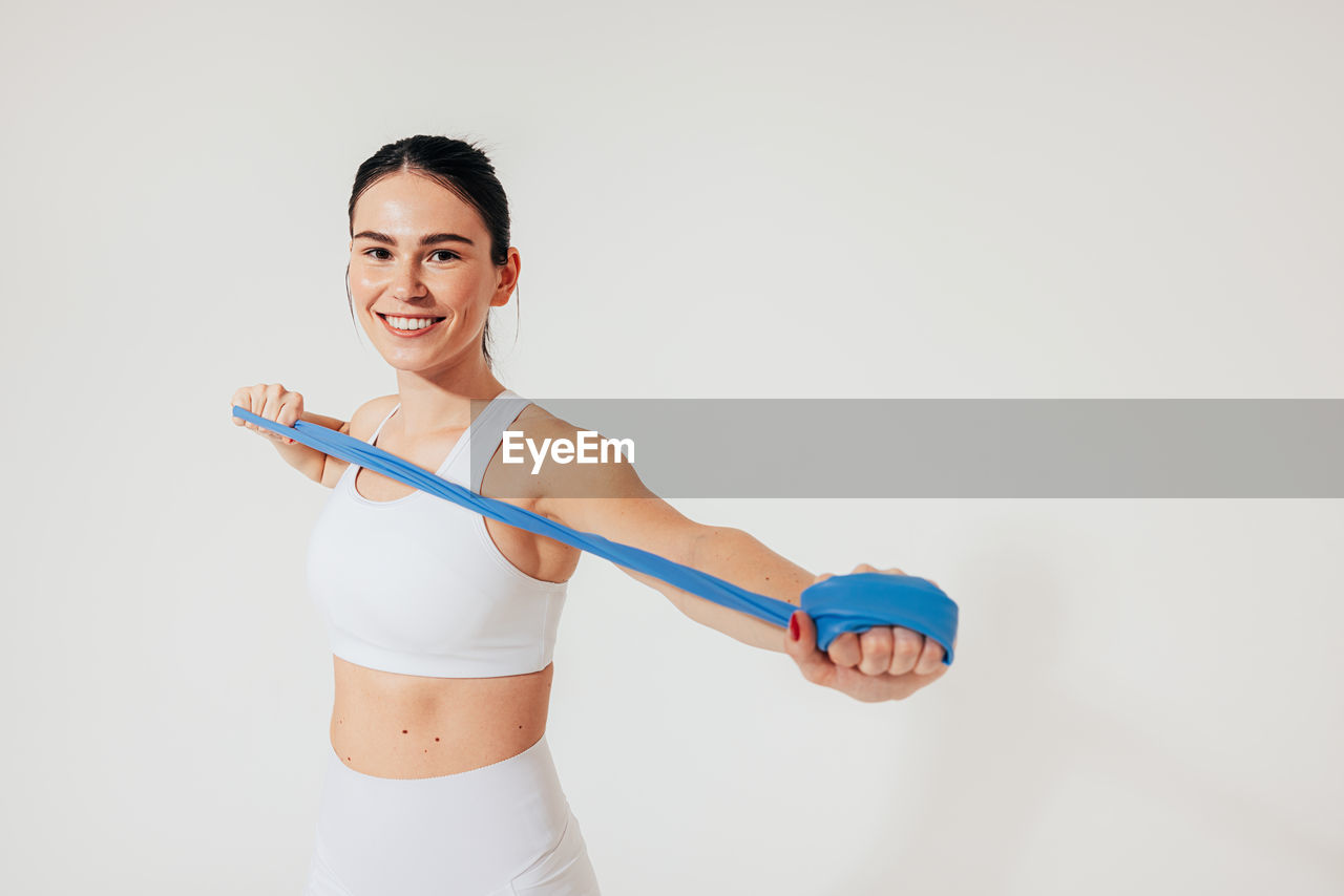 portrait of smiling young woman exercising with tape measure against white background