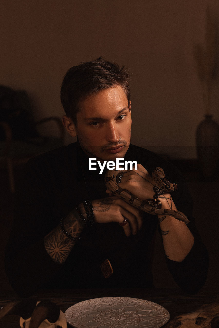 Portrait of young man holding snake sitting by table at home