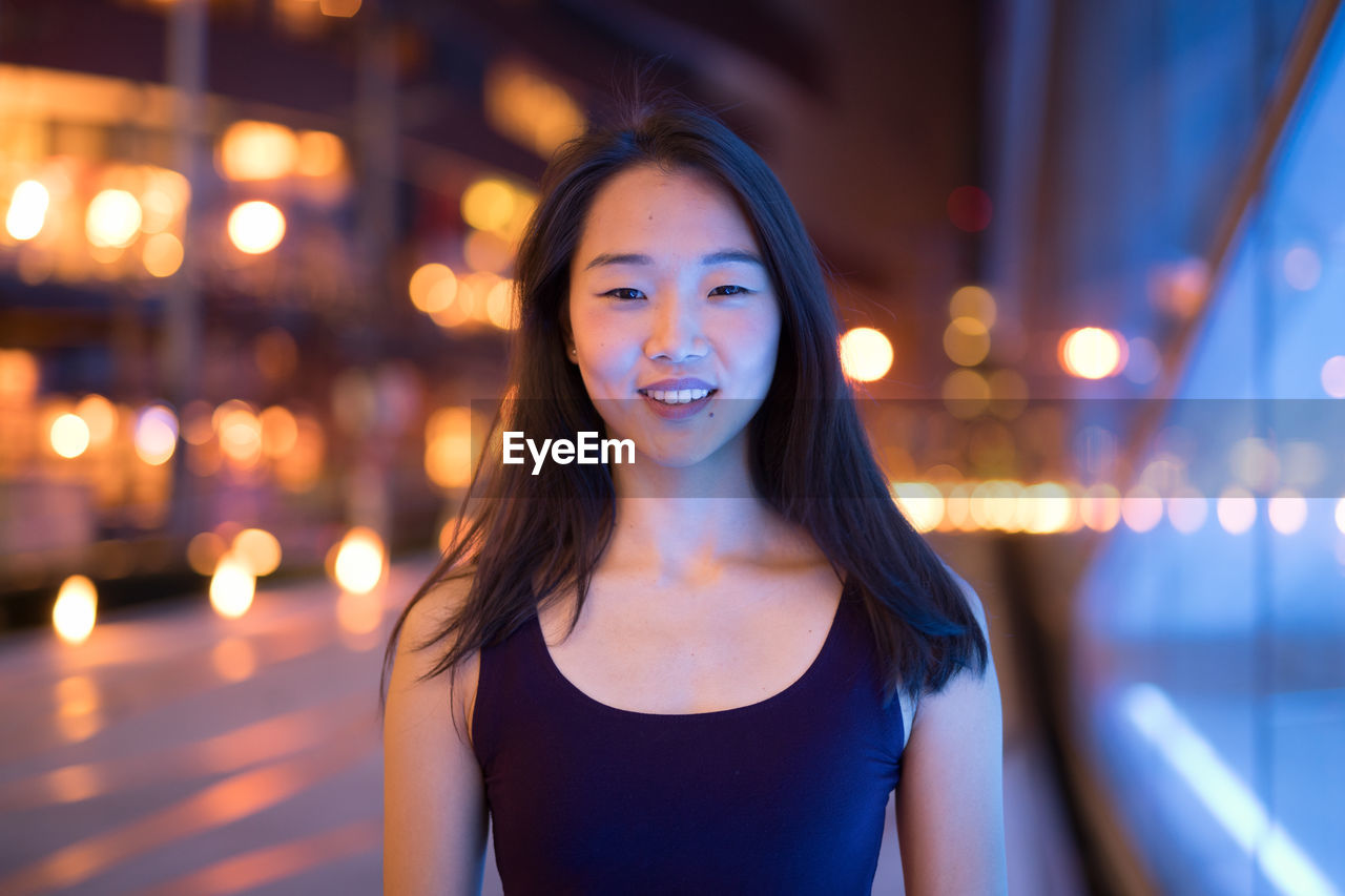 PORTRAIT OF SMILING YOUNG WOMAN STANDING AGAINST ILLUMINATED LIGHTS