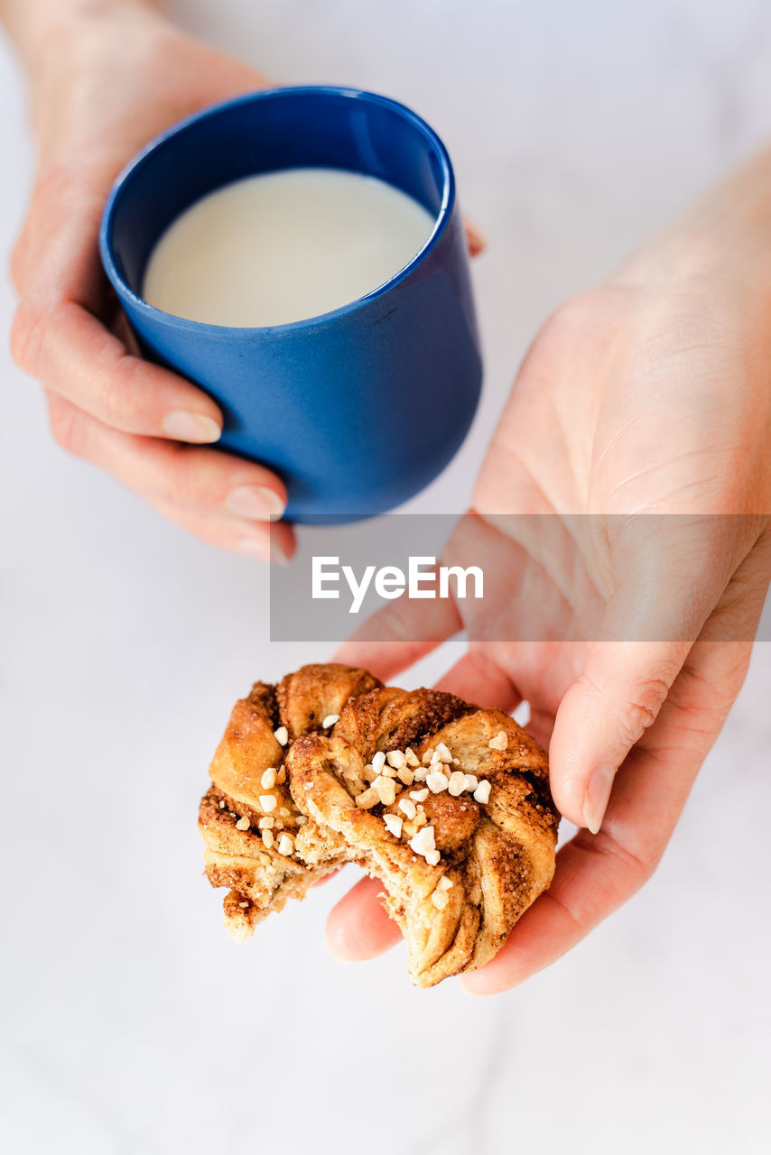 Close up of female hands holding a blue cup of milk and a cinnamon roll.