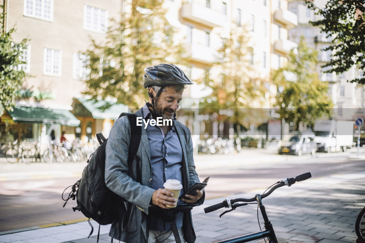 Mature businessman with disposable cup using smart phone by bicycle