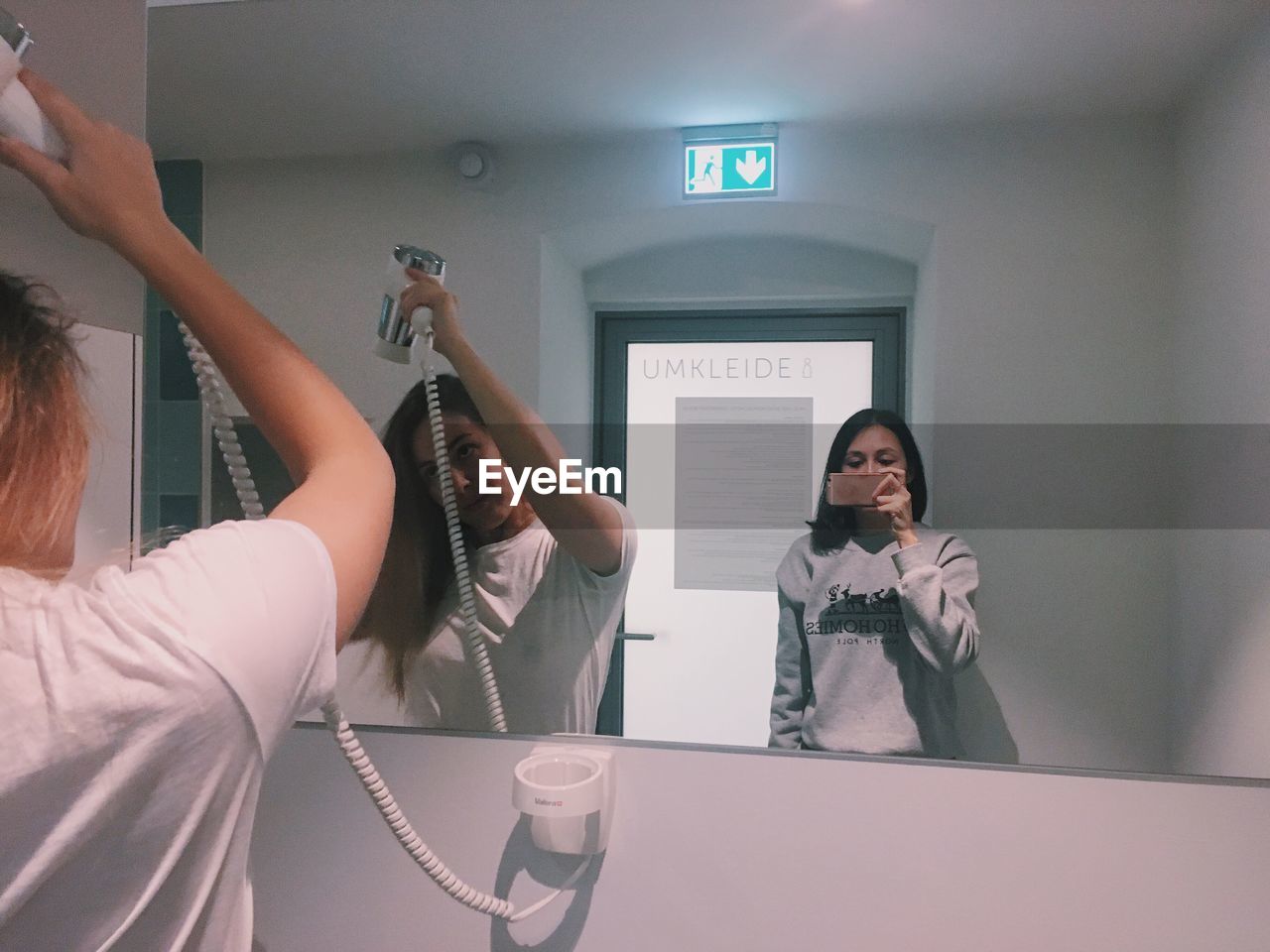Young woman taking mirror selfie while friend drying hair in restroom