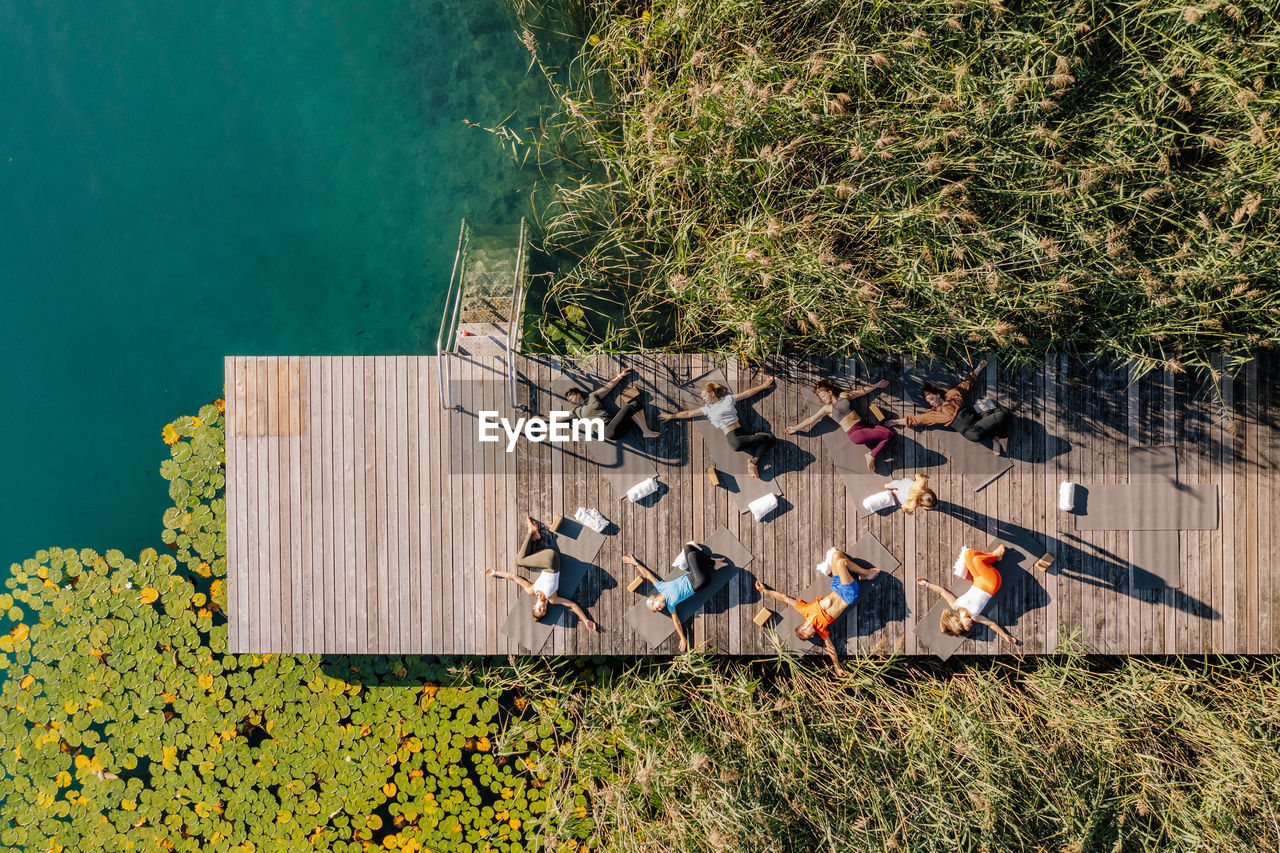 Men and women practicing yoga on jetty by lake during sunny day