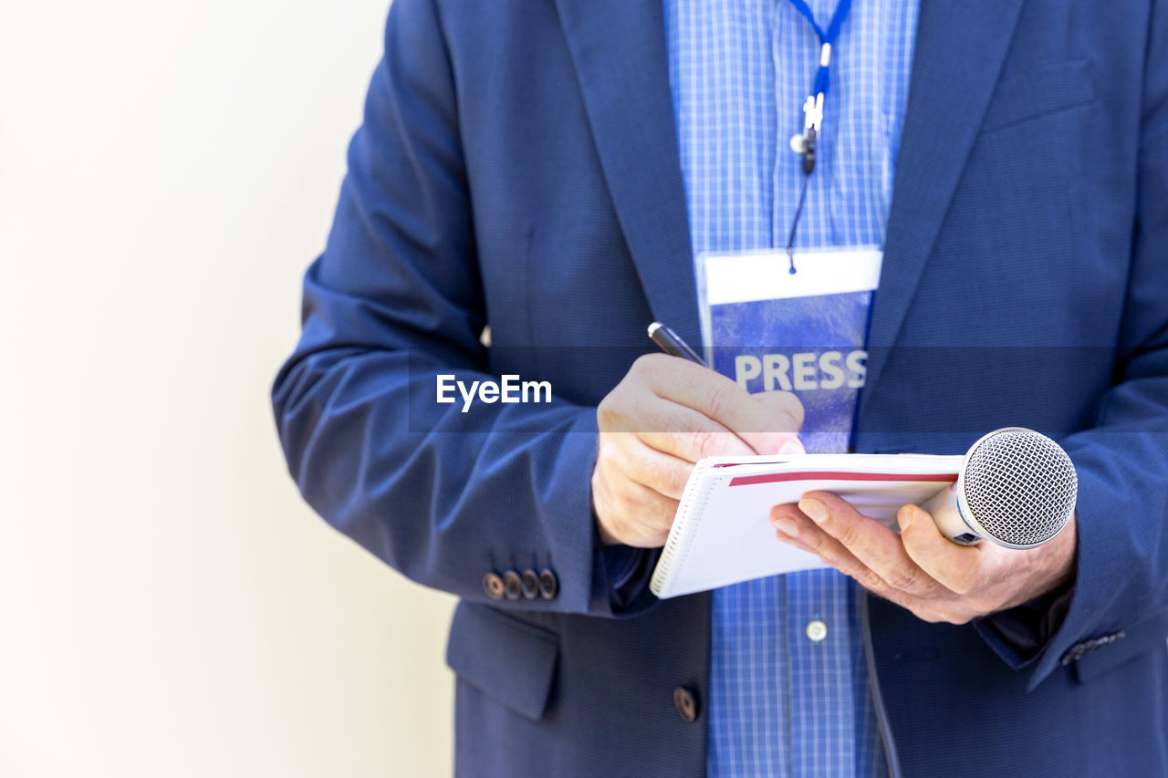 Male reporter with press pass at news conference or media event, holding microphone, writing notes