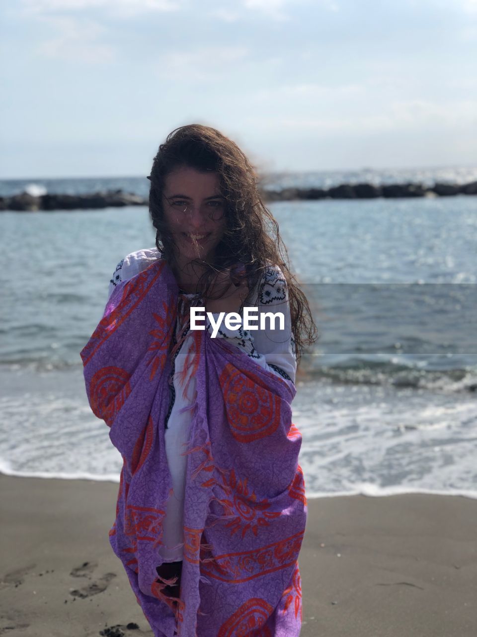 Portrait of young woman wrapped in scarf standing at beach against sky