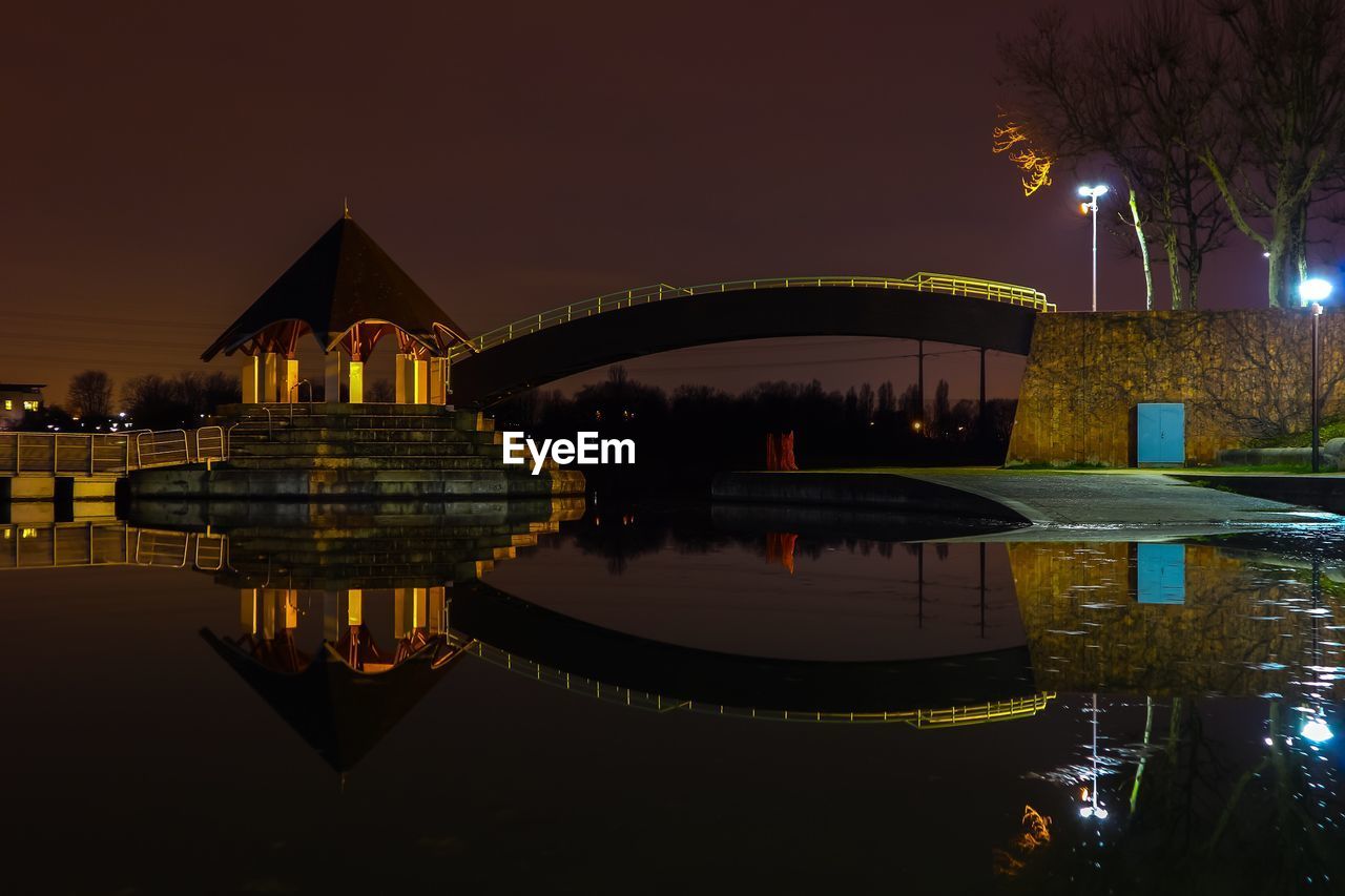 Illuminated bridge over river by buildings against sky at night