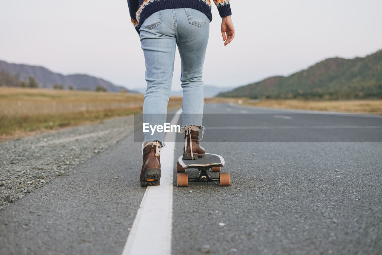 Low section of woman skateboarding on road against field