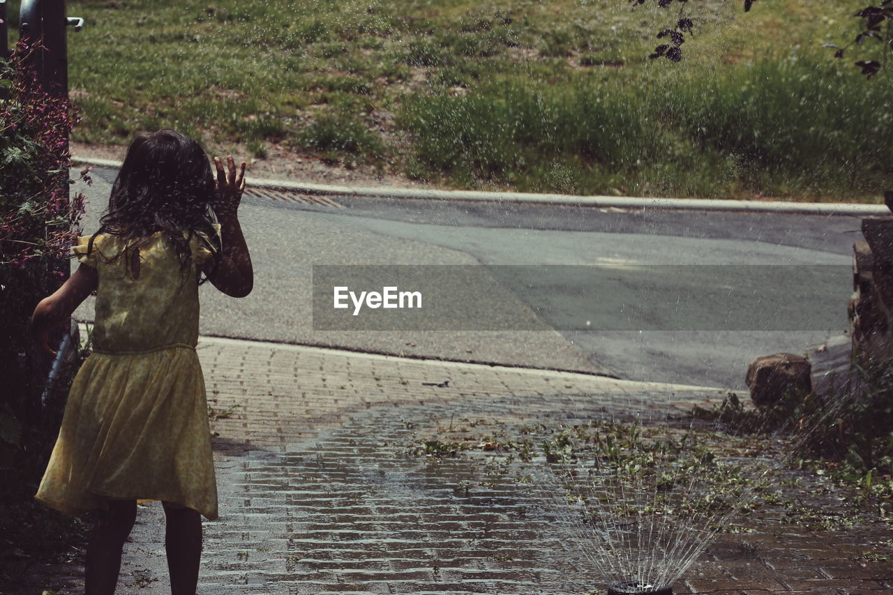 Rear view of girl standing by sprinkler against road