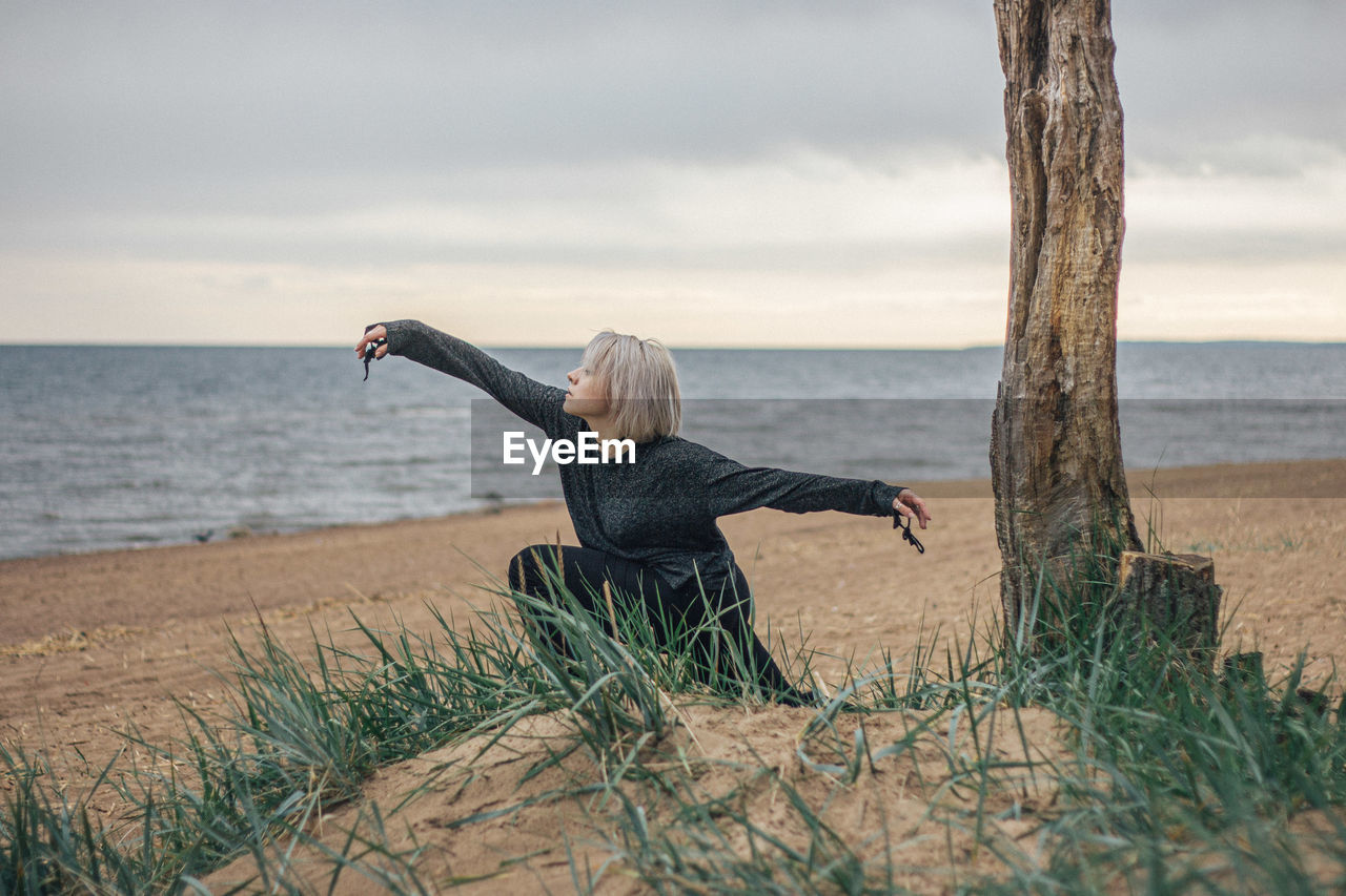 Young woman exercising while crouching by tree trunk at beach