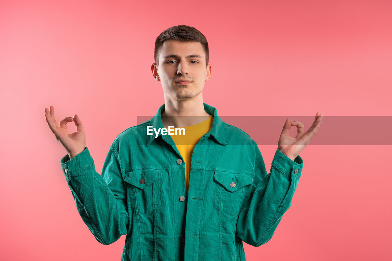 studio shot, one person, colored background, portrait, young adult, looking at camera, adult, waist up, indoors, front view, men, standing, gesturing, emotion, sleeve, pink background, clothing, outerwear, sign language, smiling, green, pink, casual clothing, happiness, arm, limb, copy space, brown hair, person, positive emotion