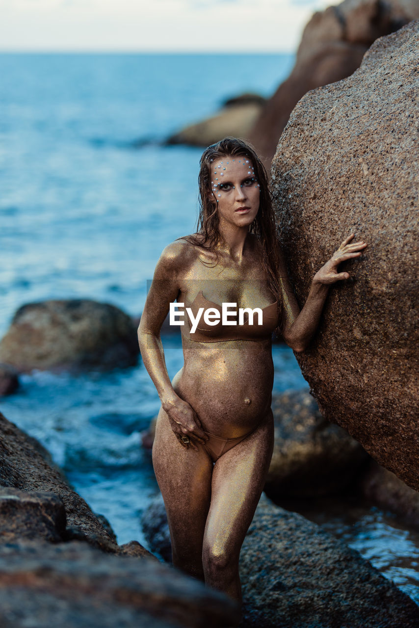 water, adult, sea, women, rock, land, one person, clothing, young adult, nature, swimwear, beach, female, bikini, portrait, photo shoot, beauty in nature, person, hairstyle, relaxation, blue, sky, holiday, long hair, vacation, trip, summer, fashion, lifestyles, outdoors, leisure activity, standing, looking at camera, day, limb, front view, full length, brown hair, human face, muscular build, sun tanning