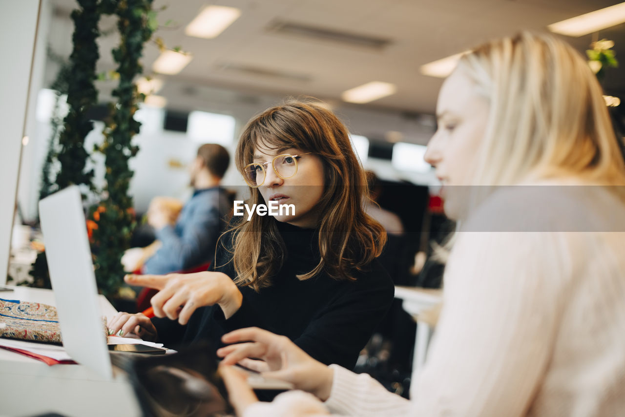 Young businesswoman pointing at laptop to female colleague sitting in office