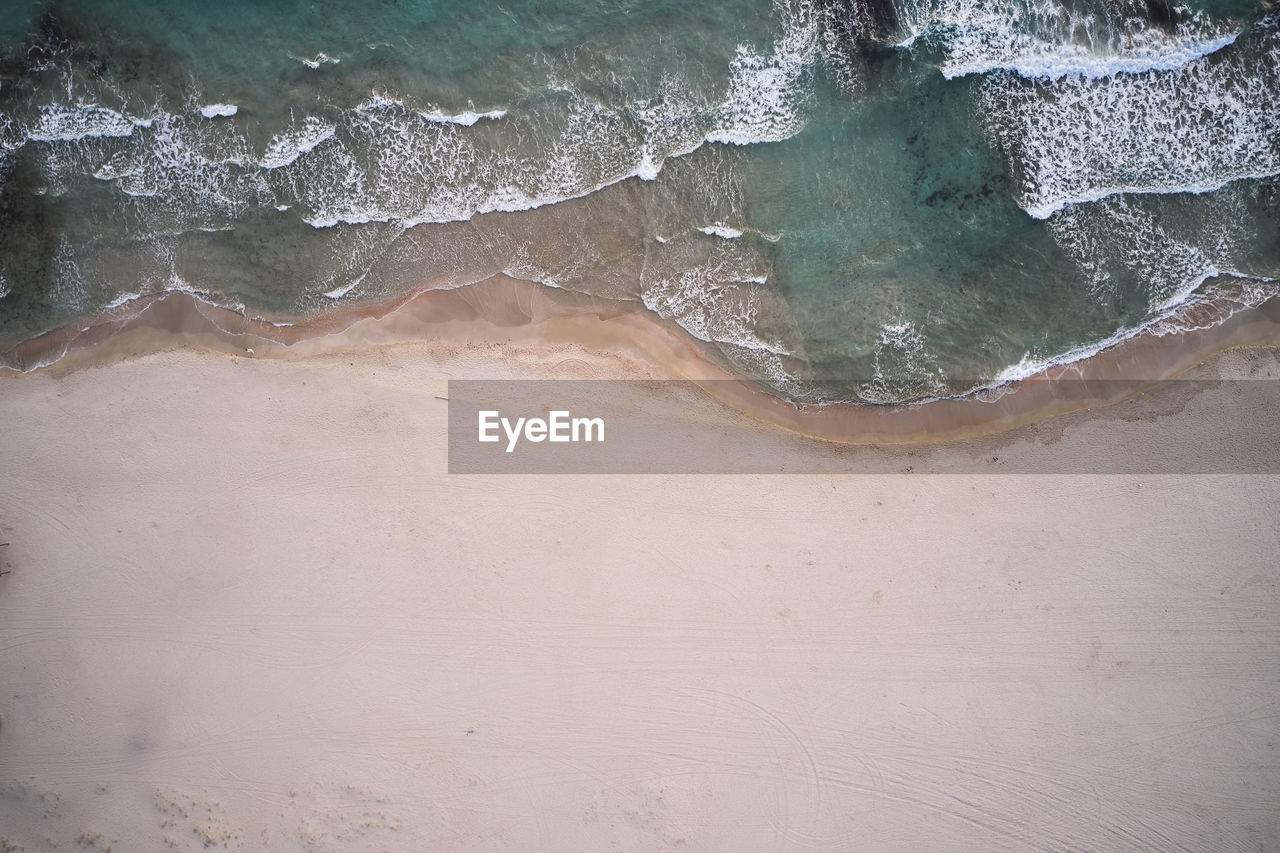 Aerial top view of turquoise water with seaweed and foamy waves rolling on empty sandy beach