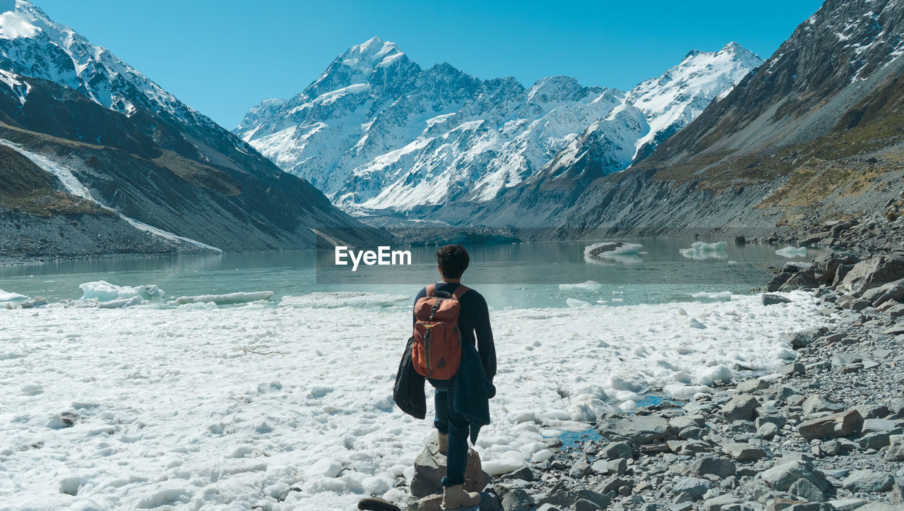 REAR VIEW OF MAN STANDING ON SNOWCAPPED MOUNTAIN