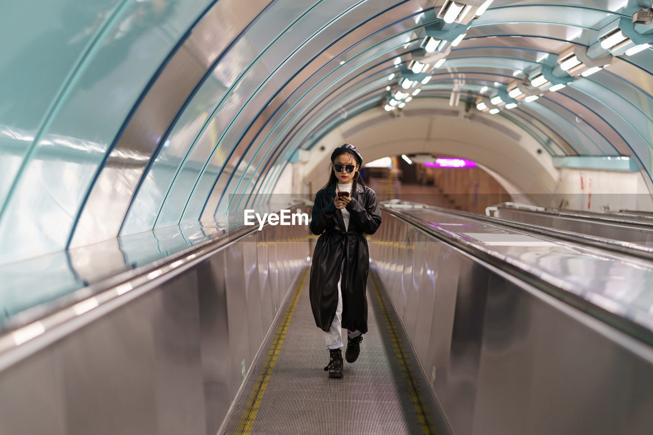 Japanese girl blogger in trendy trench and glasses on escalator use smartphone read follower comment