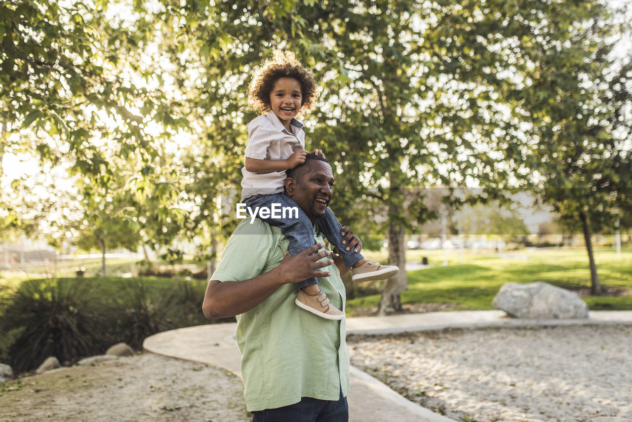 Portrait of cheerful son sitting on father's shoulders while playing at park