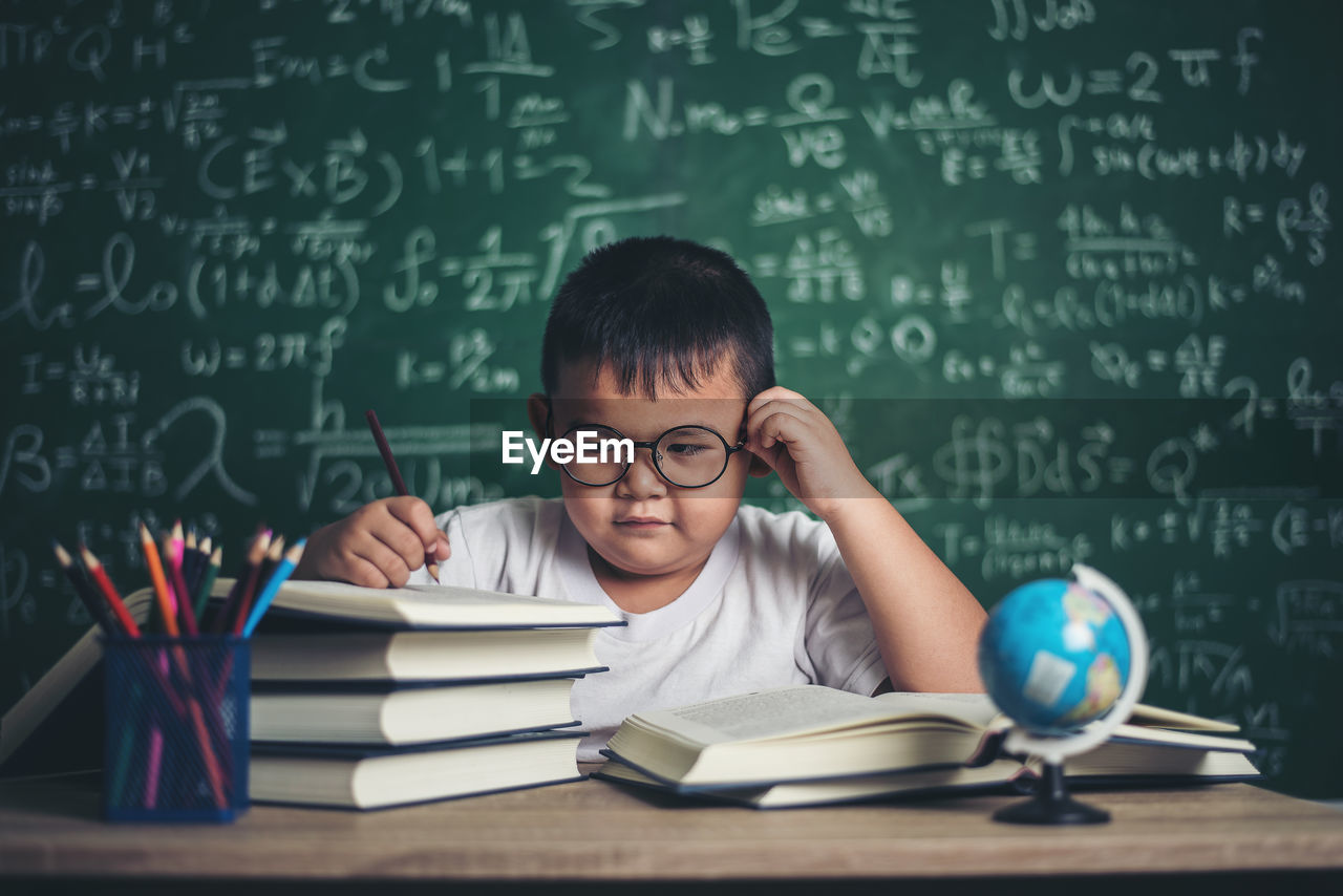 Cute boy studying at table against blackboard