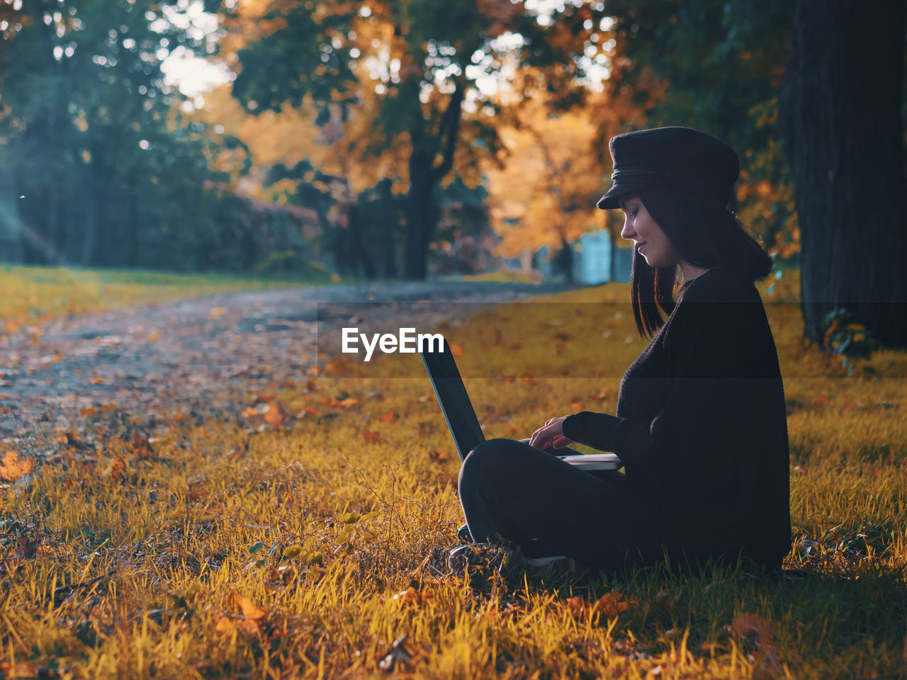 Side view of young woman using laptop while sitting at public park during autumn