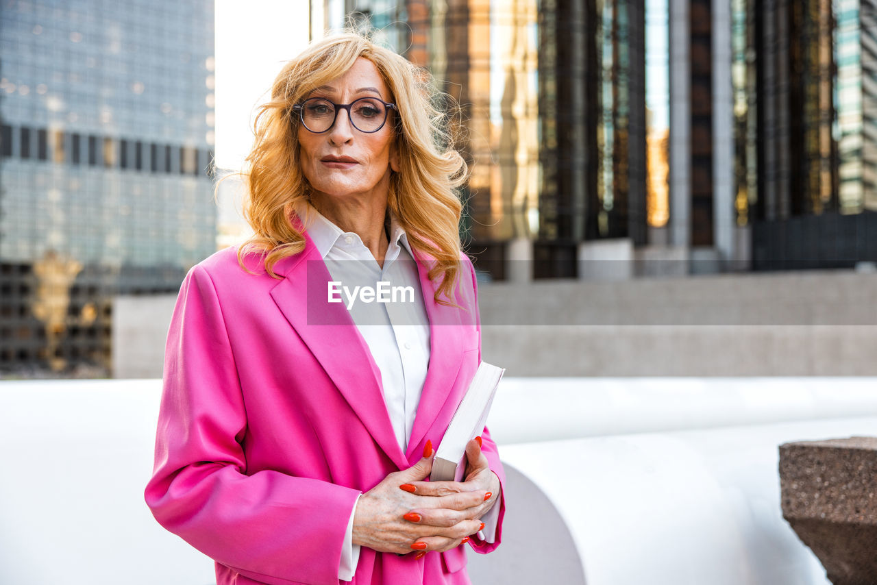 adult, women, architecture, city, one person, business, female, businesswoman, glasses, building exterior, clothing, built structure, standing, spring, city life, smiling, portrait, lifestyles, outerwear, business finance and industry, fashion, waist up, hairstyle, person, eyeglasses, happiness, young adult, professional occupation, office building exterior, communication, day, looking, mature adult, pink, outdoors, blond hair, office, technology, building, blazer, front view, copy space, wireless technology, occupation, emotion, street