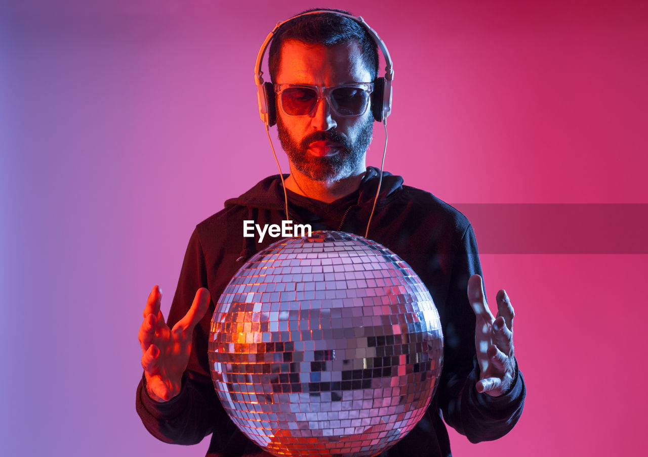 Studio portrait of a bearded deejay with headphones and sunglasses against red and blues background.