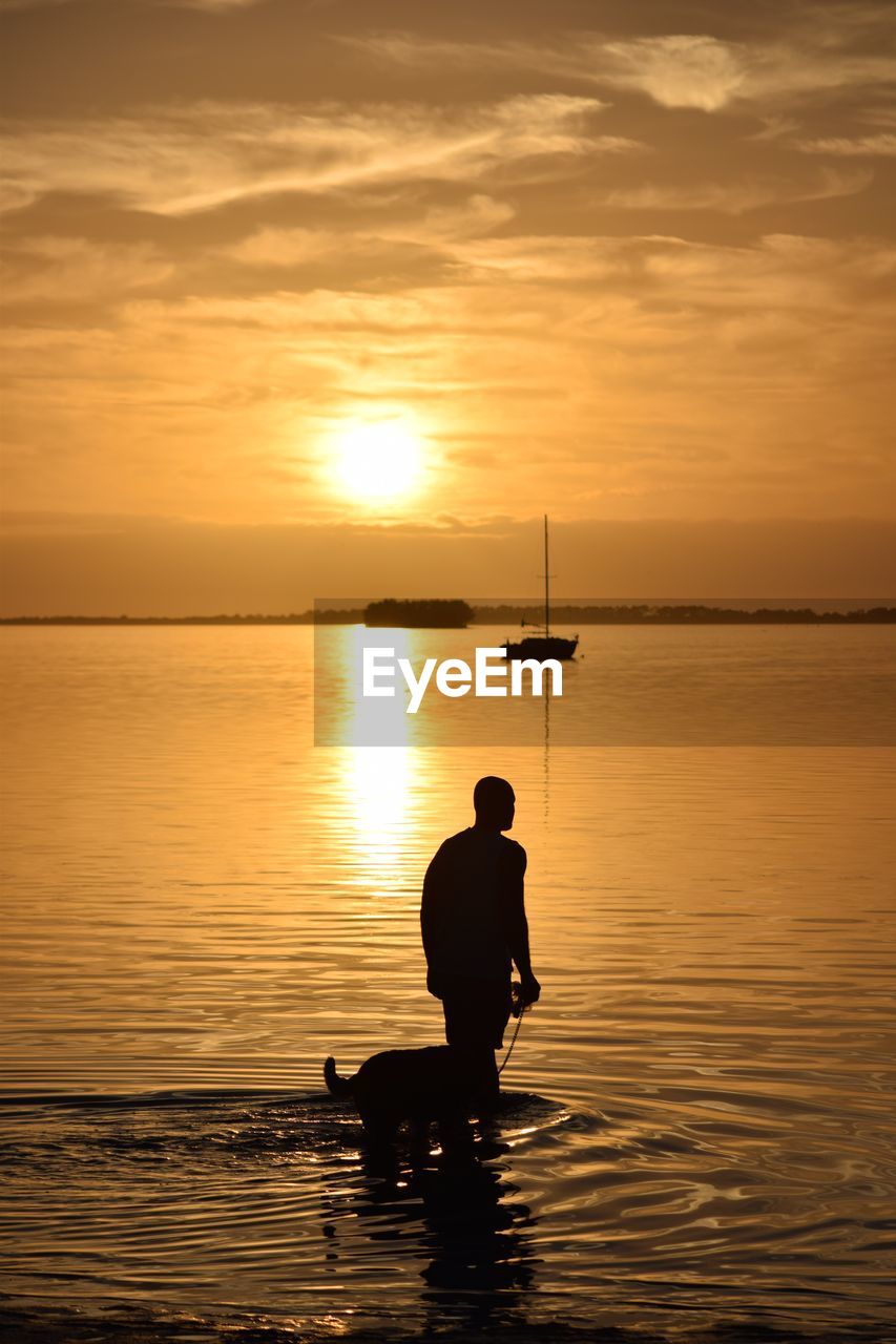 Silhouette man with dog standing in sea against sky during sunset