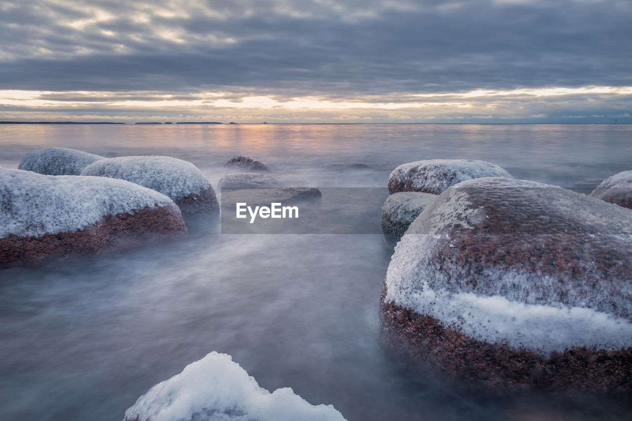 Snow covered rocks at beach against cloudy sky during sunset