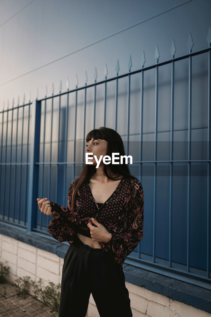 Stylish young ethnic female with long dark hair in trendy dress standing against blue wall on street and looking away thoughtfully