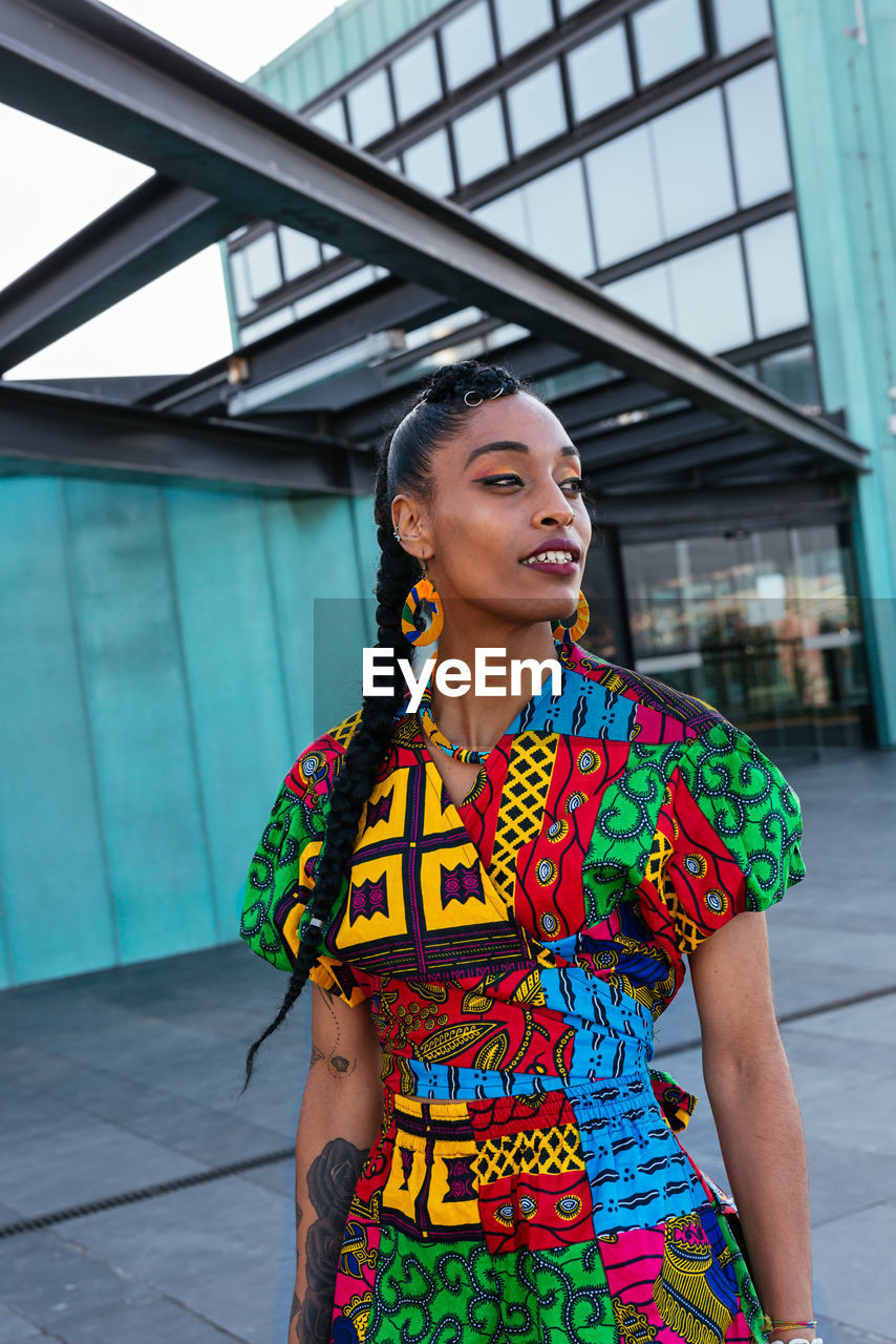 Young ethnic female in braids in colorful clothes looking away while standing near turquoise wall of apartment building in city