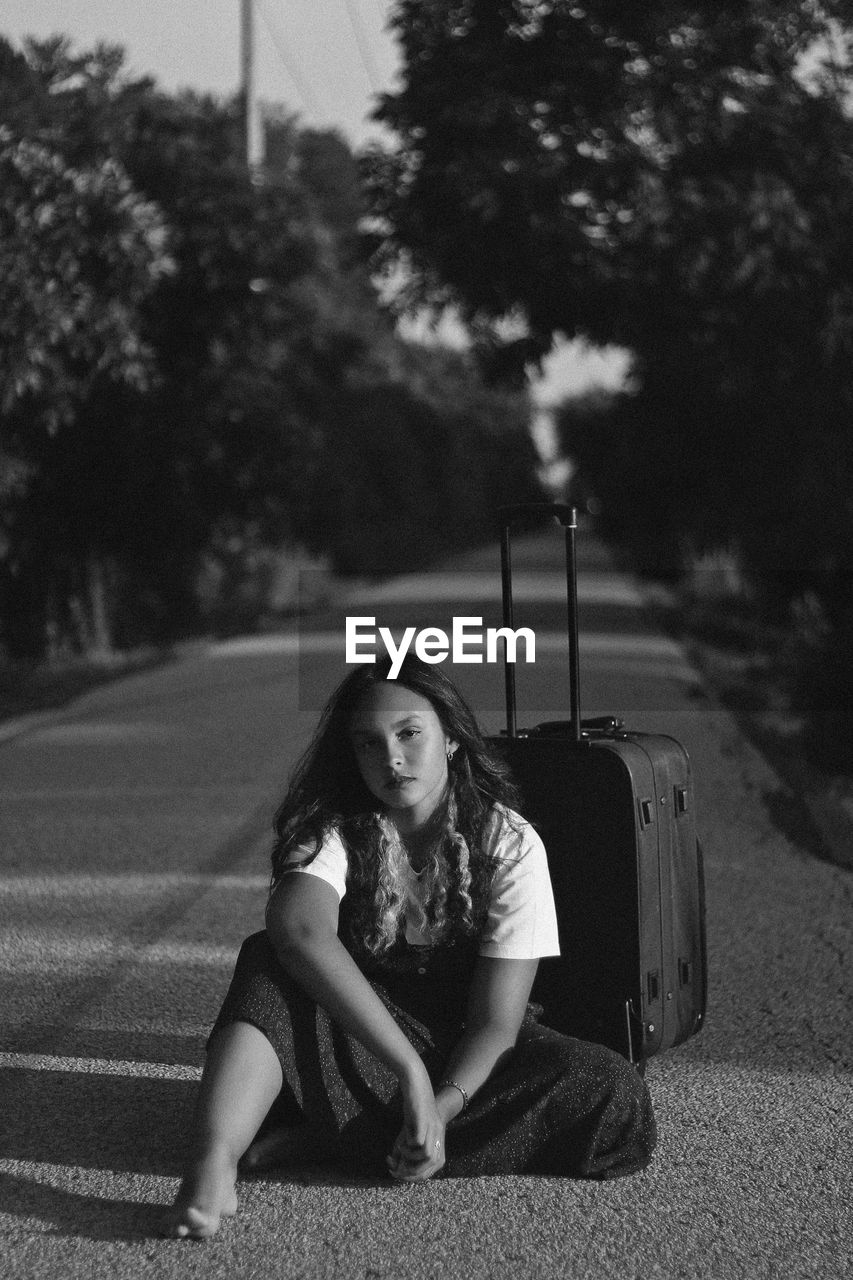 Portrait of young woman sitting with luggage on road