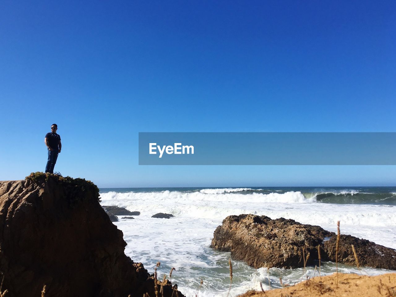 Man standing on rock by sea against clear blue sky