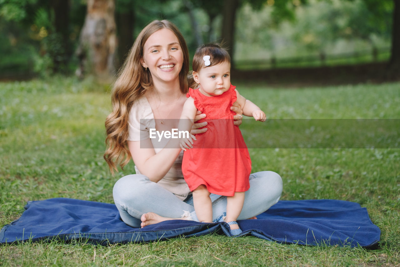 MOTHER AND DAUGHTER SITTING ON GRASS WHILE LOOKING AT FARM