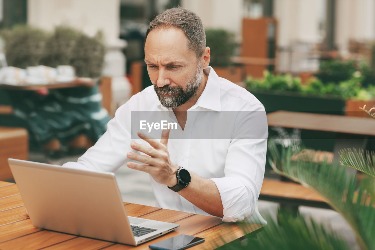 An adult man sits on the street in a cafe, communicates on the network and works on a laptop.